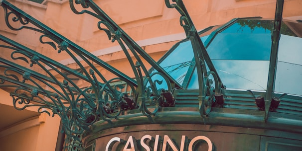 The image displays the entrance to a casino, characterized by detailed and ornate architectural elements with green metal structures and a sign that reads 'CASINO'. The background includes a glass dome and a peach-colored wall.