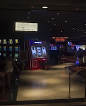 A dimly lit casino with several rows of slot machines, some displaying bright graphics and lights. There are a few people scattered throughout the area, engaging with the machines. The floor is reflective and polished, adding to the ambiance, while the ceiling is dotted with numerous small lights.