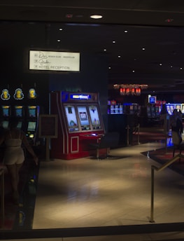 A dimly lit casino with several rows of slot machines, some displaying bright graphics and lights. There are a few people scattered throughout the area, engaging with the machines. The floor is reflective and polished, adding to the ambiance, while the ceiling is dotted with numerous small lights.