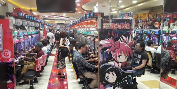 A bustling arcade filled with people playing various gaming machines. Brightly colored displays and animated characters are prominent on posters and screens. The atmosphere is lively with patrons seated in rows, focusing on the games. Overhead signage and electronic scoreboards are visible, contributing to the busy and vibrant environment.