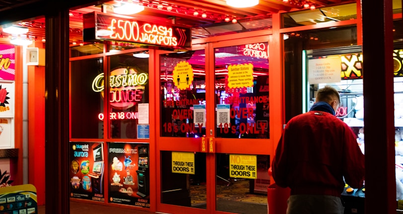 A brightly lit entrance to a casino with neon signs, including one advertising a £500 jackpot. The doors have clear signage indicating entry is restricted to those over 18. A person in a red jacket stands to the right, possibly interacting with a machine or display.