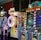 A row of brightly colored arcade game machines is arranged in a line against the wall. Each machine features vibrant lights and designs, with themes ranging from claw games to game show-inspired setups. The area has a playful and inviting atmosphere, suitable for entertainment or amusement centers.