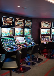 A row of brightly lit slot machines in a casino setting, each with colorful displays and various game themes. The machines are lined up against a dark wall and are unoccupied, with cushioned chairs placed in front of them. The area is dimly lit except for the glow from the screens, creating a vibrant and engaging atmosphere.