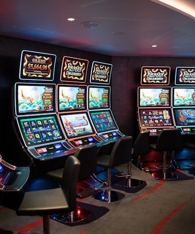 A row of brightly lit slot machines in a casino setting, each with colorful displays and various game themes. The machines are lined up against a dark wall and are unoccupied, with cushioned chairs placed in front of them. The area is dimly lit except for the glow from the screens, creating a vibrant and engaging atmosphere.