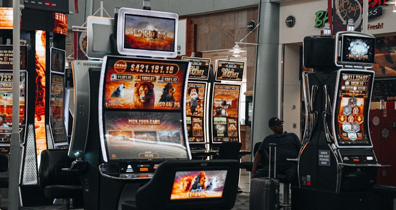 A collection of slot machines is situated inside a building with bright, colorful screens displaying various games and gaming themes. A person is seated nearby, seemingly relaxed with luggage beside them, and the atmosphere is lively with electronic displays.